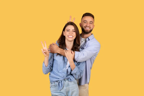 Laughing woman flashing peace sign with playful man making bunny ears behind her head, both enjoying light-hearted moment on yellow background