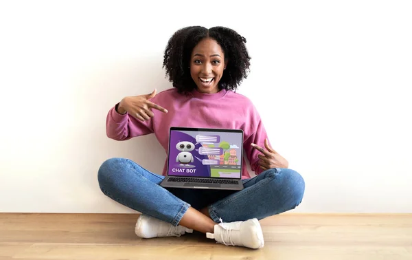 Excited young black woman pointing at laptop with chatbot on screen. Happy millennial african american lady sitting on floor over white blank wall in empty apartment, recommending AI assistant