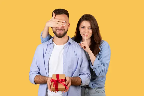 Woman covers mans eyes for surprise gift moment, her finger to her lips for secrecy, as he holds gift with red ribbon, on yellow background