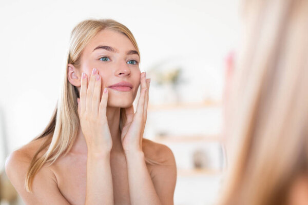 Pretty blonde woman applying moisturizer to her face gently touching cheeks, enjoying pampering skincare ritual looking at mirror reflection in cozy bathroom setting. Selective focus
