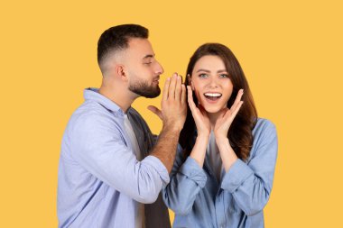 Elleri yanaklarında neşeli bir kadın, mavi giyinmiş, sarı arka planda neşeli bir anı paylaşan, fısıldayan bir adamı dinler.