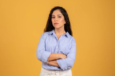 Kendine güvenen, uzun kıvırcık saçlı, kollarını kavuşturmuş açık mavi bir gömlek giyen, güçlü sarı bir arka plana karşı duran güçlü ve kendine güvenen genç bir kadın.