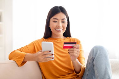 Kredi kartı ve akıllı telefonuyla çekici genç Asyalı kadın evdeki koltukta oturuyor, bankacılık ya da internetten alışveriş yapıyor, mobil uygulama kullanıyor, kapatıyor.