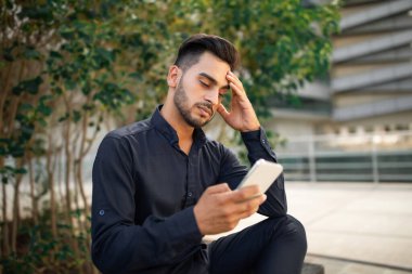 Genç Arap iş adamı akıllı telefondan kıyamet kopuyor, internette kötü haberleri okuyor ve mesajlaşıyor, dışarıda ofis arkaplanında otururken stresi gösteriyor. İş yerinde sorunlar, iletişim sorunları