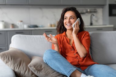 Bluz ve günlük kot giymiş neşeli genç bir kadın telefonunda iç açıcı sohbetlerden hoşlanır. Gri koltukta rahatça oturur, şık bir oturma odasında.