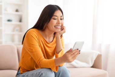 Uzun saçlı mutlu Asyalı kadın kanepede oturuyor, telefonda haberler okuyor, online video içeriği izliyor, oturma odası içi. Çinli kadın hafta sonunun keyfini çıkarıyor, akıllı telefondan kayıyor.