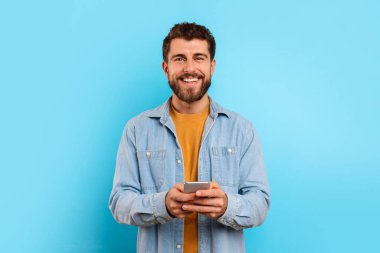 Akıllı telefonlu neşeli adam kameraya bakıyor ve gülümsüyor, sosyal medyada mavi stüdyo geçmişi üzerinden mesaj atıyor. Telefonla sohbet eden, yeni mobil uygulama kullanan pozitif genç adam.