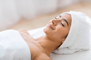 Huzurlu, çekici, havluya sarılı genç bir Hintli kadın. Kapalı gözlerle rahatlıyor ve lüks spa merkezindeki tüm vücut tedavi prosedürlerinden sonra gülümsüyor..