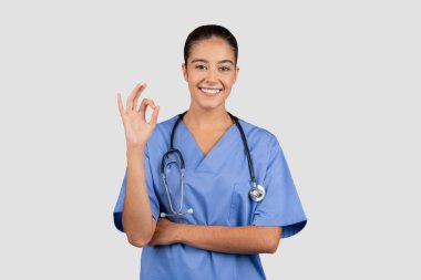 Bin yıllık olumlu beyaz kadın doktor ya da hemşire. Mavi üniformalı. Gri stüdyo arka planında izole edilmiş el yazısı var. Profesyonel sağlık hizmeti onayı, satış tavsiyesi