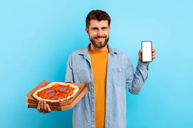 Pizza kutusu olan aç genç adam mavi stüdyo arka planında duran akıllı telefon boş ekranını gösteriyor. Adam abur cubur teslimatı için pizzacı başvurusu öneriyor. Model