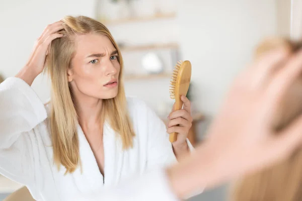 Young blonde woman with concern examines her hair for dandruff, utilizing a brush for gentle care in her modern bathroom setting, showing signs of distress over hair loss. Selective focus