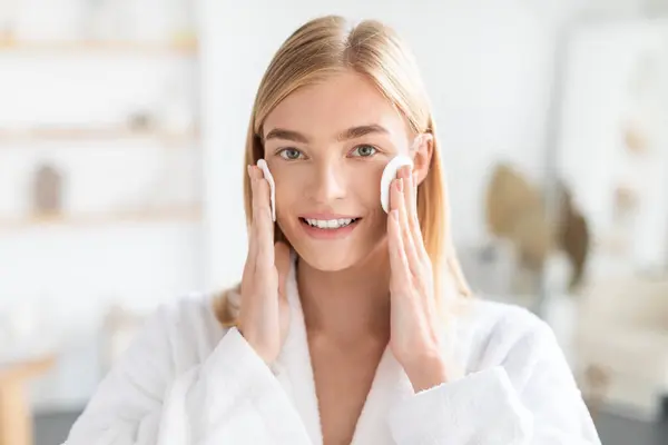 Skincare cosmetics. Pretty blonde lady finds joy in selfcare holding cotton pads against her face, standing in light modern bathroom, radiating natural beauty and wellness