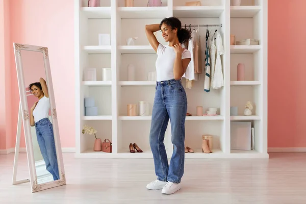 Smiling latin woman feeling confident in her high-waisted jeans, striking pose while looking at her reflection in full-length mirror in pink room