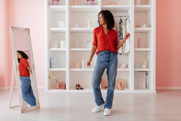 Relaxed black woman enjoying her outfit, red blouse paired with blue jeans, checking her reflection in full-length mirror in a pastel room.
