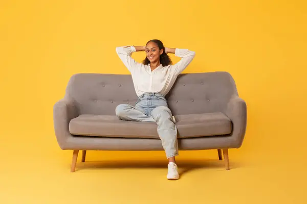 Relaxed black teenager girl in casual wear resting sitting on comfortable couch against yellow studio backdrop. Teen lady enjoys free time and relaxation, full length shot