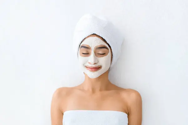 Face mask, spa beauty treatment. Top view of joyful young indian woman with white facial clay mask on her face resting on table at spa salon, skincare concept, white background, copy space