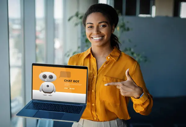 Cheerful millennial black woman standing at office, pointing at computer screen, using chatbot on laptop, collage. African american lady using artificial intelligence technologies in business