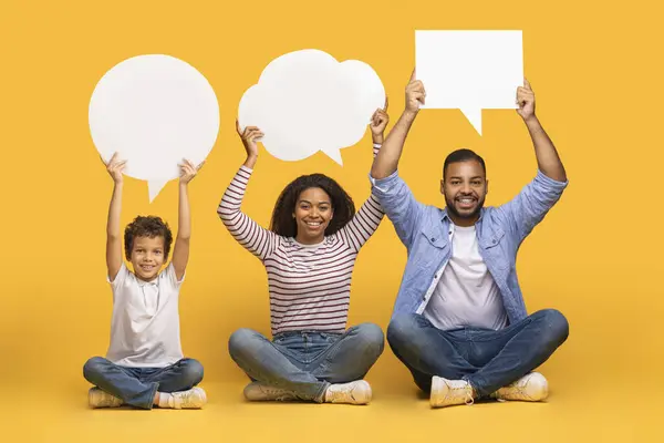 Diverse Opinions. Black Mom, Dad And Little Son Holding Different Empty Speech Bubbles Above Their Heads, African American Family Of Three Sitting On Floor On Yellow Studio Background, Copy Space