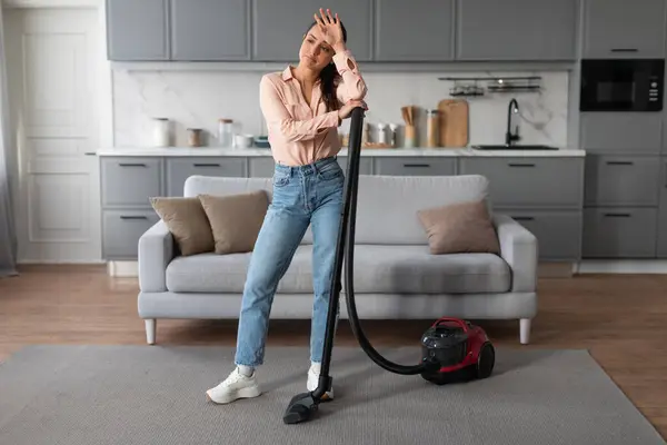 Feeling fatigued, woman takes moment to rest her head on her hand while vacuuming, reflecting brief respite amid her household duties