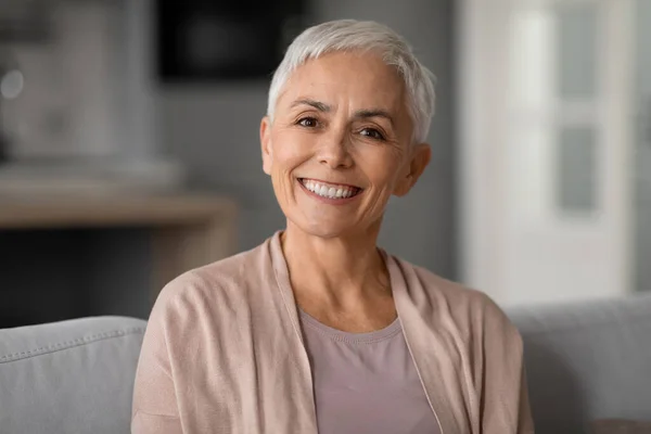 Portrait of happy healthy older woman with short haircut and gray hair posing for photo at home, smiling to camera with cheerful expression, sitting on comfortable couch at home.