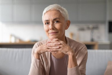 Kendine güvenen, şık saç kesimi olan, el ele tutuşup kameraya sakin bir şekilde bakan, modern iç mekanda koltukta oturan, kendine güvenen son sınıf Avrupalı kadının portresi.