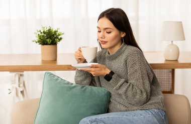 Evdeki kanepede oturan, kahve içen, tembel hafta sonunun tadını çıkaran esmer, rahat bir kadın. Mutlu bayan aromatik bitkisel çay kokulu, fotokopi alanı