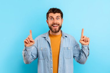 Kot gömlekli heyecanlı genç adam parmaklarını kafasının üstündeki fotokopi alanını işaret ederek mavi arka planda iki eliyle harika bir teklif sunuyor, pozitif enerji yayıyor.