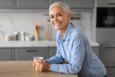 Gülümseyen son sınıf öğrencisi kadın masanın yanında durup sabah kahvesinin tadını çıkarıyor, modern yerli mutfağında elinde fincanla poz veriyor, mutlu bir ifadeyle kameraya bakıyor. Sıcak içecek içen kadın portresi