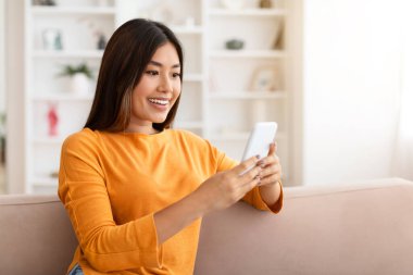 Mutlu çekici Çinli kadın elinde telefonla kanepede dinleniyor, kayıyor, web sörfü yapıyor, güzel online teklifleri kontrol ediyor, anlaşma yapıyor, mobil uygulama kullanıyor, ev içini kullanıyor, kopyalama alanı kullanıyor.
