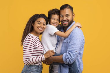 Şefkatli siyahi aile, ortada oğlu, grup kucaklaşması, neşeli Afro-Amerikan ailesi ve erkek çocuk mutluluk saçıyor, sarı stüdyo geçmişine karşı birlikte poz veriyorlar.
