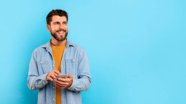 Gülümseyen genç adam cep telefonuyla mesajlaşıyor ve yan bakıyor, mavi stüdyonun arka planında boş alanın yanında duruyor, panorama. Mutlu milenyum çocuğu akıllı telefondan uygulamalara bakıyor.