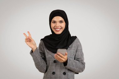 Tesettürlü, tesettürlü, gülümseyen Müslüman iş kadını, elinde akıllı bir telefonla modern bir bağlantı ve pozitiflik gösterirken barış işareti yapıyor.