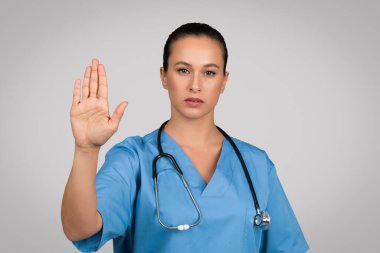 Mavi üniformalı ciddi bir Avrupalı kadın doktor dur işareti yapıyor, bir şeyi reddediyor, virüslere hayır diyor, gri arka planda tek başına duruyor.
