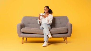 Bir fincan sıcak kahveli mutlu siyah kız sarı stüdyo arkaplanına karşı rahat koltukta oturuyor, tek başına dinlenirken lezzetli içkinin tadını çıkarıyor. Tam boy çekim, panorama