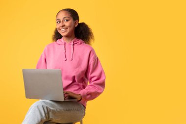 Bilgisayarlı siyah genç kız internette öğreniyor sarı arka planda oturuyor, dijital yerli öğrencinin internet teknolojisiyle çalıştığı bir stüdyo fotoğrafı. E-öğrenme teklifleri, kopyalama alanı
