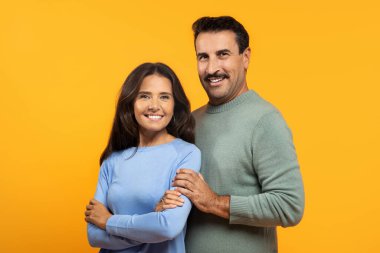 Mutlu, yaşlı, beyaz bir çift, erkeğin kolları kadının etrafında, neşeli turuncu bir arka planda, kameraya doğru güvenle gülümsüyor.
