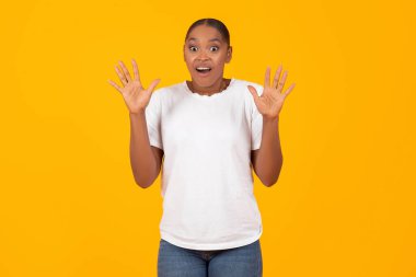 Şok olmuş genç bir kadın, sarı stüdyo arka planında dikilip korkmuş bir ifadeyle kameraya bakarken elleri havada poz verirken yakalandı. Ben değilim, suç kavramı.