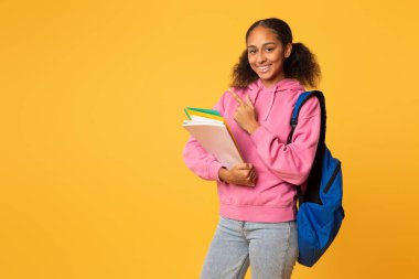 Neşeli afro-amerikan öğrenci kolej kitaplarını ve defterleri tutuyor, mavi sırt çantasıyla poz veriyor ve sarı stüdyo arka planına karşı boş alanı işaret ediyor. Eğitim teklifi