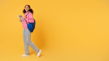 Eğitim Teklifi. Afrika kökenli Amerikalı genç kız üniversite kitaplarını elinde tutuyor mavi sırt çantasıyla, sarı stüdyo arka planına karşı mesaj atmak için boş alanın yakınında duruyor. Panorama
