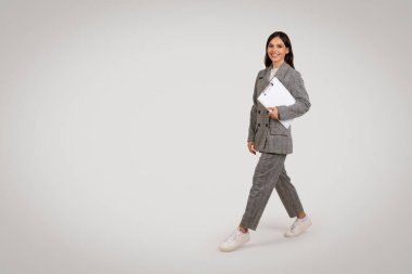 Dinamik genç profesyonel elinde dizüstü bilgisayarla yürüyor, modaya uygun ekoseli takım elbiseler giyiyor, gündelik spor ayakkabılarla eşleşiyor, modern iş kıyafetlerini somutlaştırıyor, tam boy