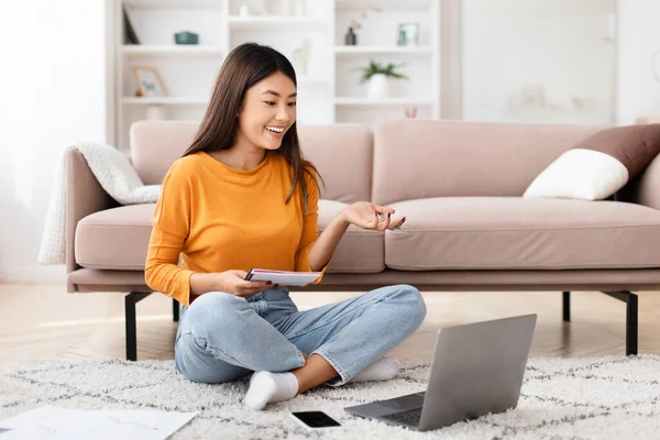 Young asian woman entrepreneur working from home, sitting on floor in living room, holding notepad, looking at laptop computer screen, gesturing, have online meeting with business partner, copy space