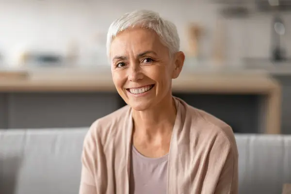 Portrait Of Smiling Senior Lady With Gray Short Hair Wearing Elegant Beige Homewear, Sitting On Couch At Home. Happy Woman Posing Indoors Expressing Positive Emotions, Headshot