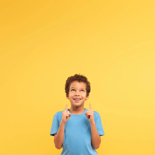 上を見上げて指で指を指差し 青いシャツを黄色の正方形の背景に期待して微笑み 楽観主義と未来を象徴する若い男の子 — ストック写真