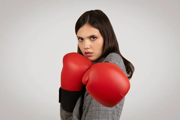 Determined young woman in business attire, poised with red boxing gloves, exuding strong defensive stance and readiness to tackle challenges head-on