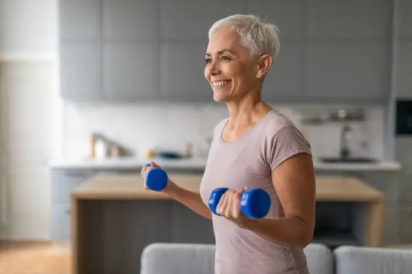 Sporty mature lady lifts blue dumbbells maintaining fitness in modern home interior, leading active sporty lifestyle and keeping fit in retirement, enjoying domestic exercises