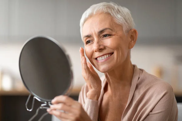Beautiful senior woman with stylish haircut looking in round mirror at home, gently touching her face and smiling, happy about perfect skin condition and anti aging skincare. Beauty care concept