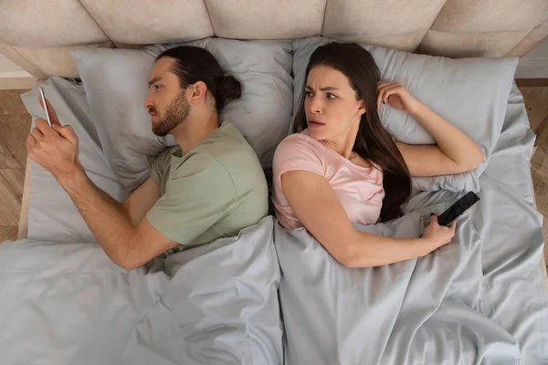 悲しい若い女性は寝室のインテリア トップビューでベッドでスマートフォンでチャットする夫に目を向けます デバイス中毒 仮想反逆 コミュニケーションリモート — ストック写真