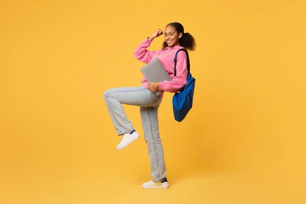 Happy black teen student lady with laptop gesturing yes while posing with backpack over yellow studio background. Full length shot of teenager learner celebrating educational success. Free space
