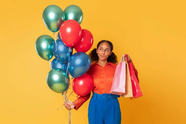 Holiday sales. Thoughtful african american teenager girl holds paper shopping bags and bunch of colorful balloons standing over yellow studio backdrop, thinking about great festive commerce offers
