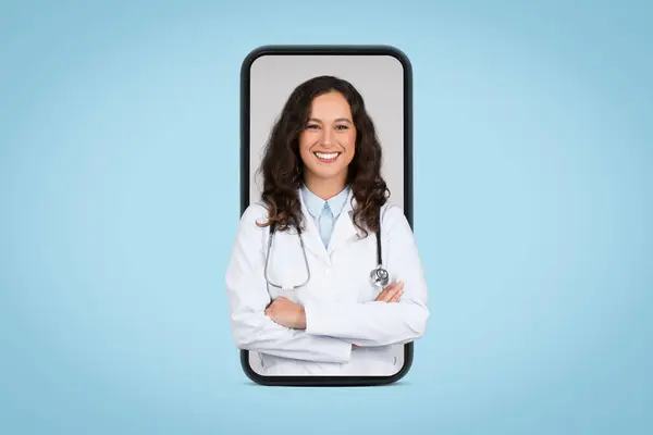 stock image Cheerful young woman doctor in white uniform posing with stethoscope and folded arms in giant cellphone screen, blue background, banner